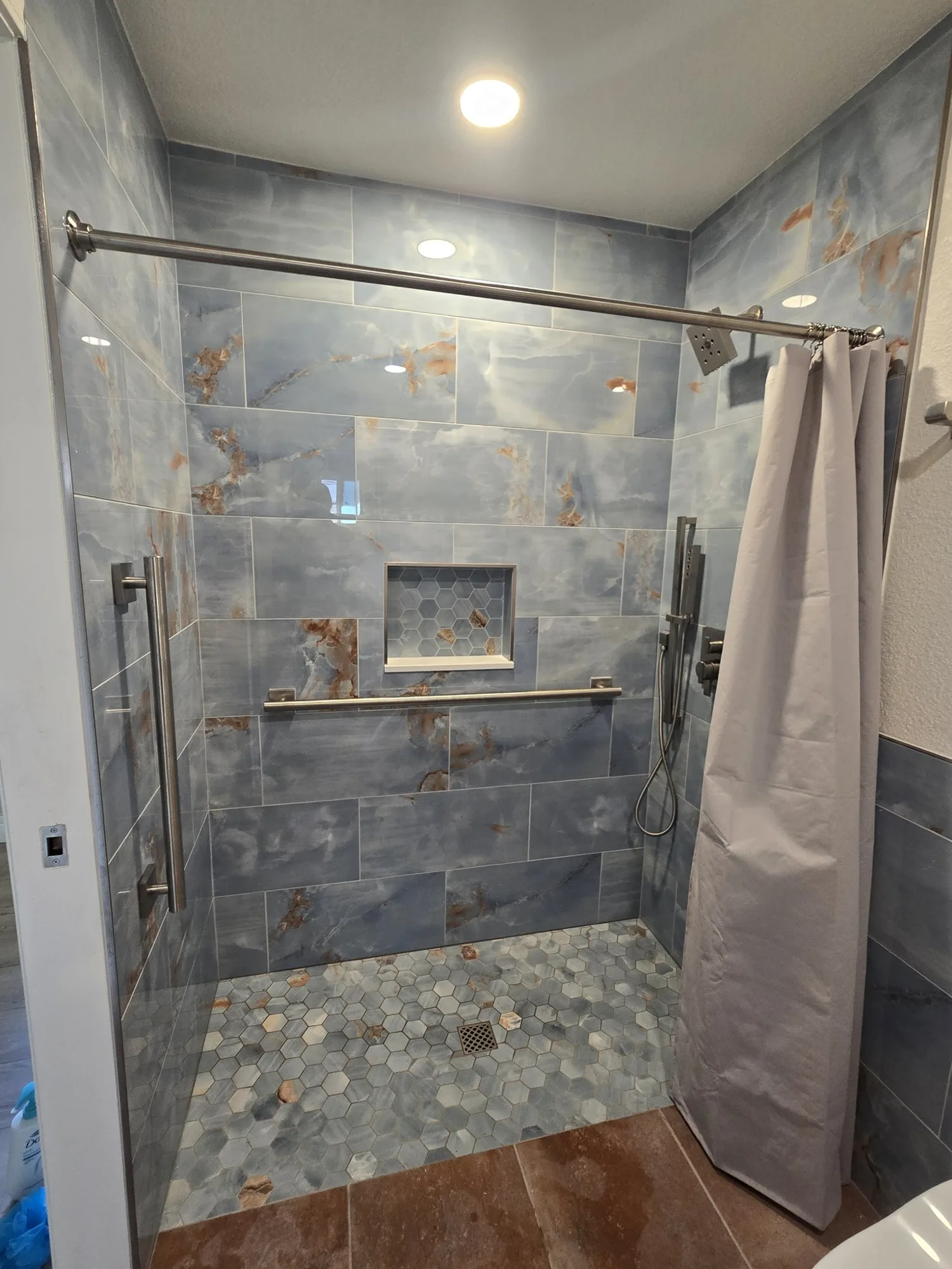 Curbless shower with blue marble-look tile, pebble floor, niche and grab bars