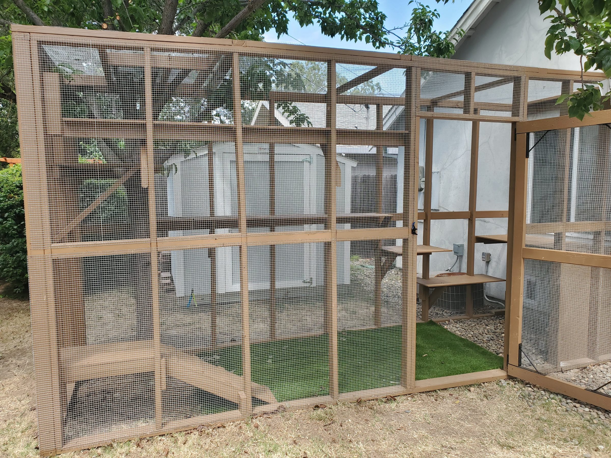 Wood-framed screened cat enclosure beside a house, with artificial turf and climbing shelves