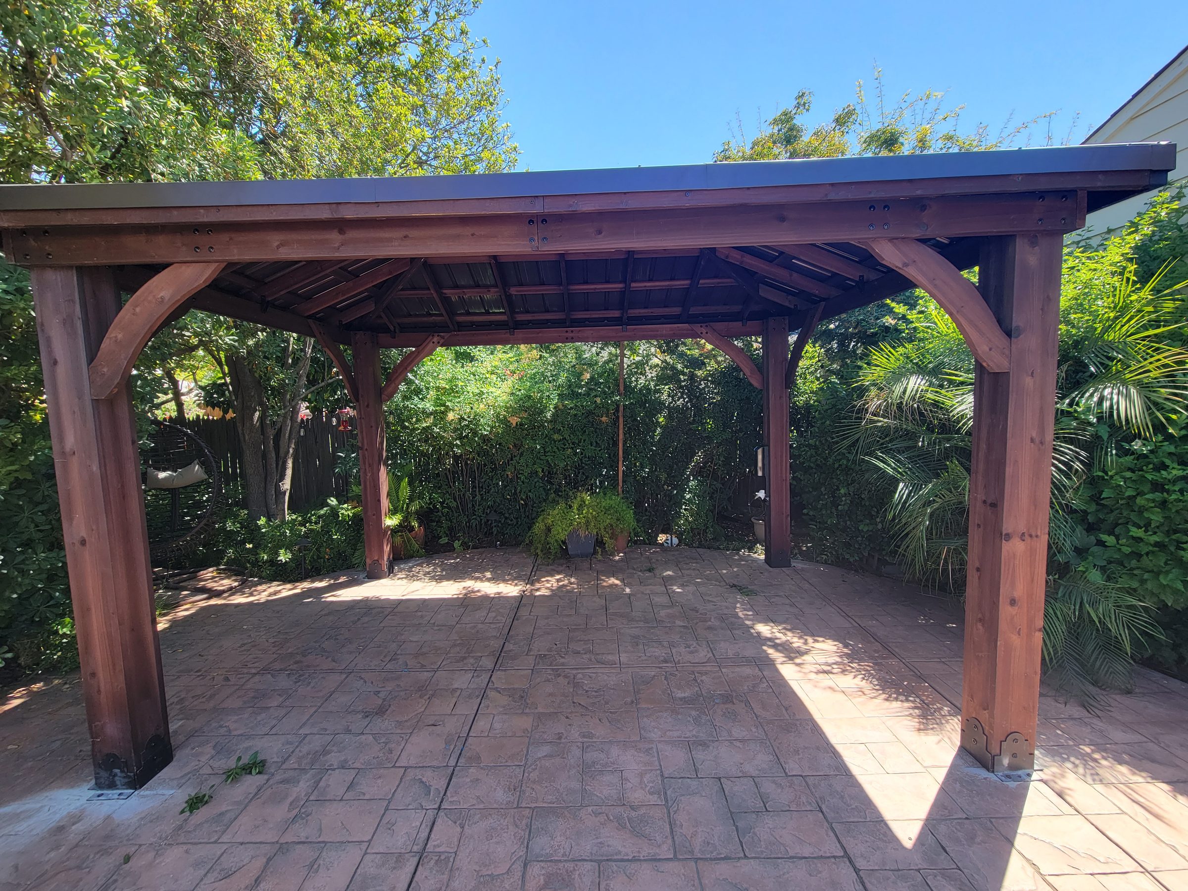 Freestanding wood gazebo with a metal roof on a stamped-concrete patio, backyard greenery behind