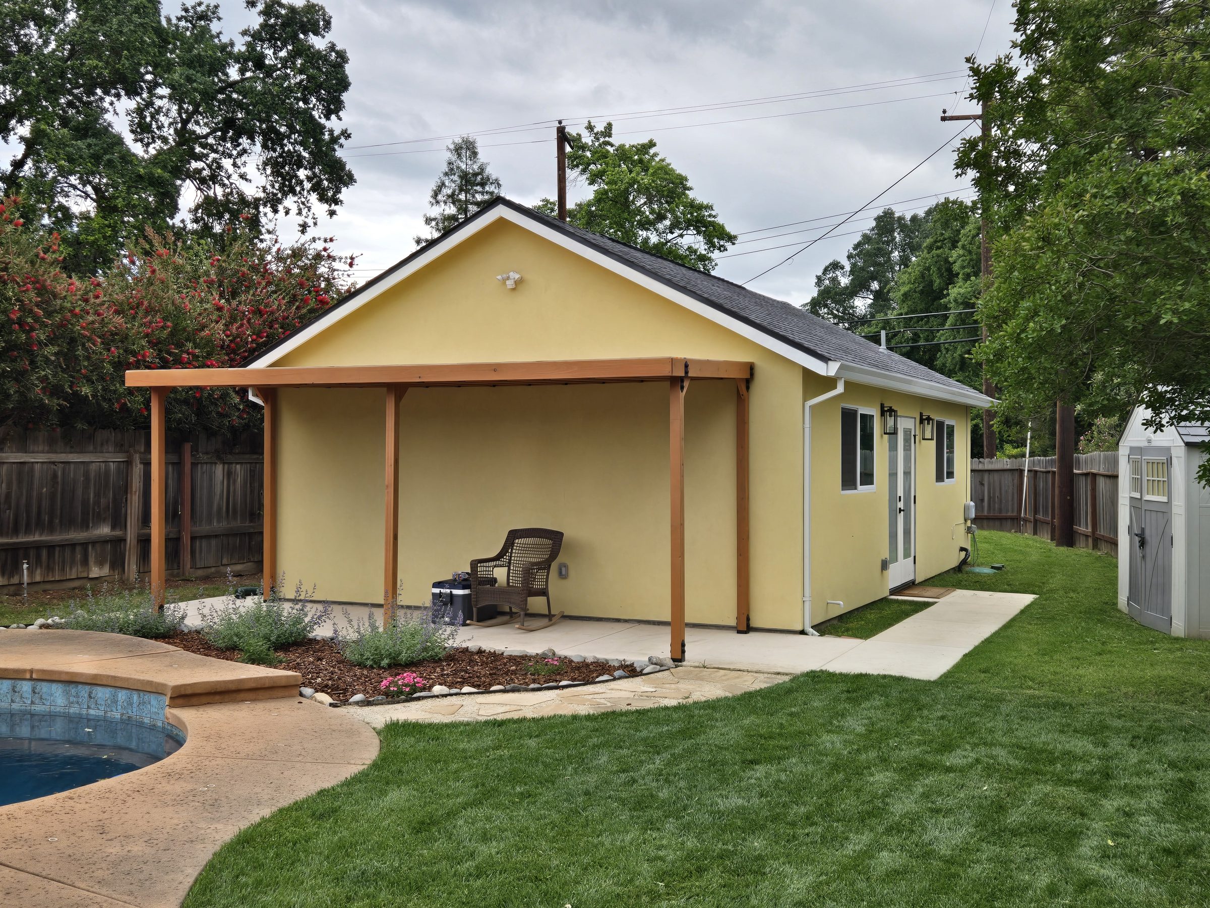 Wood-framed covered patio along the side of a yellow stucco building, with a pool and lawn beside it