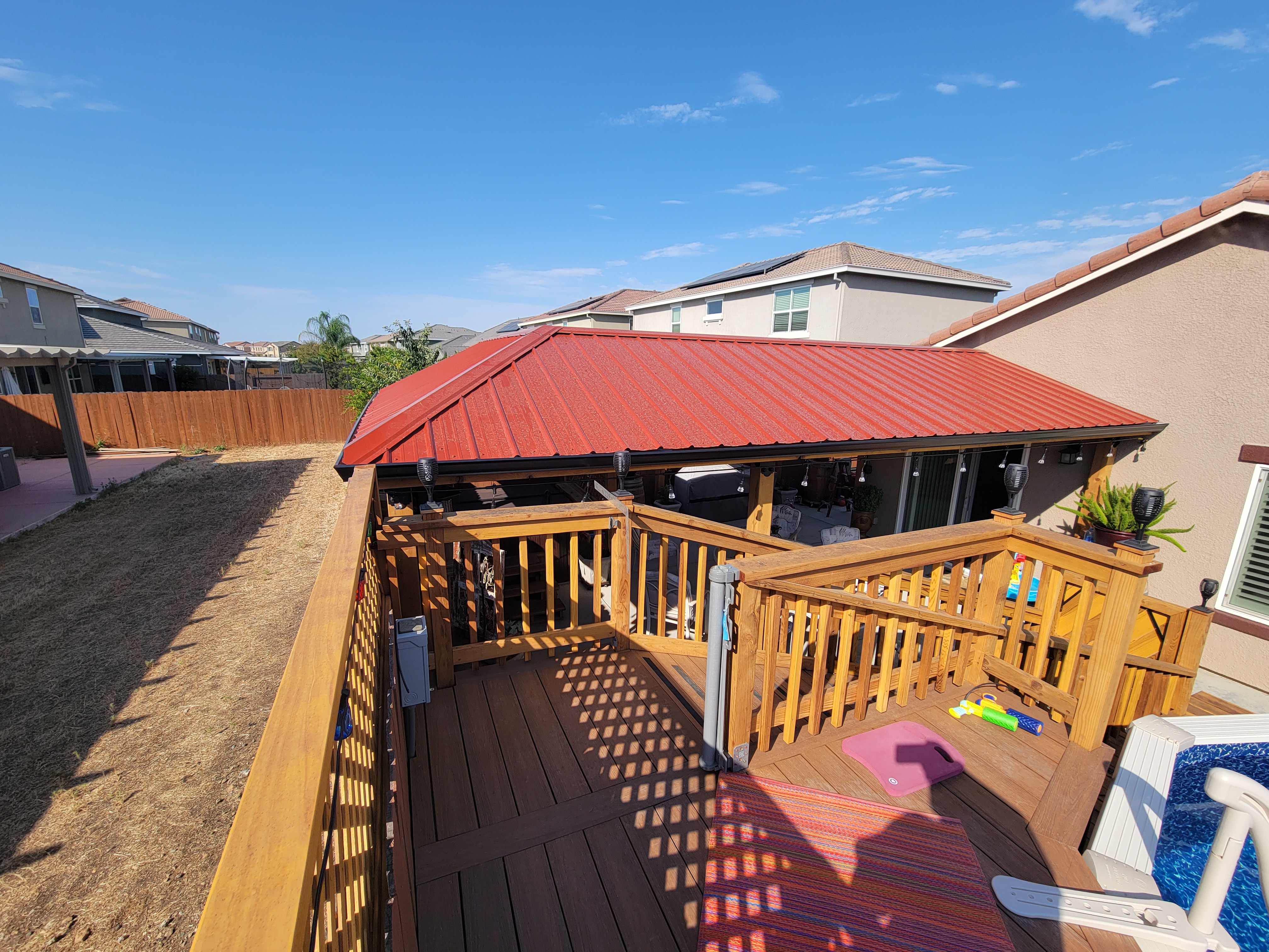 Covered patio with a red standing-seam metal roof over a wood deck beside a backyard pool