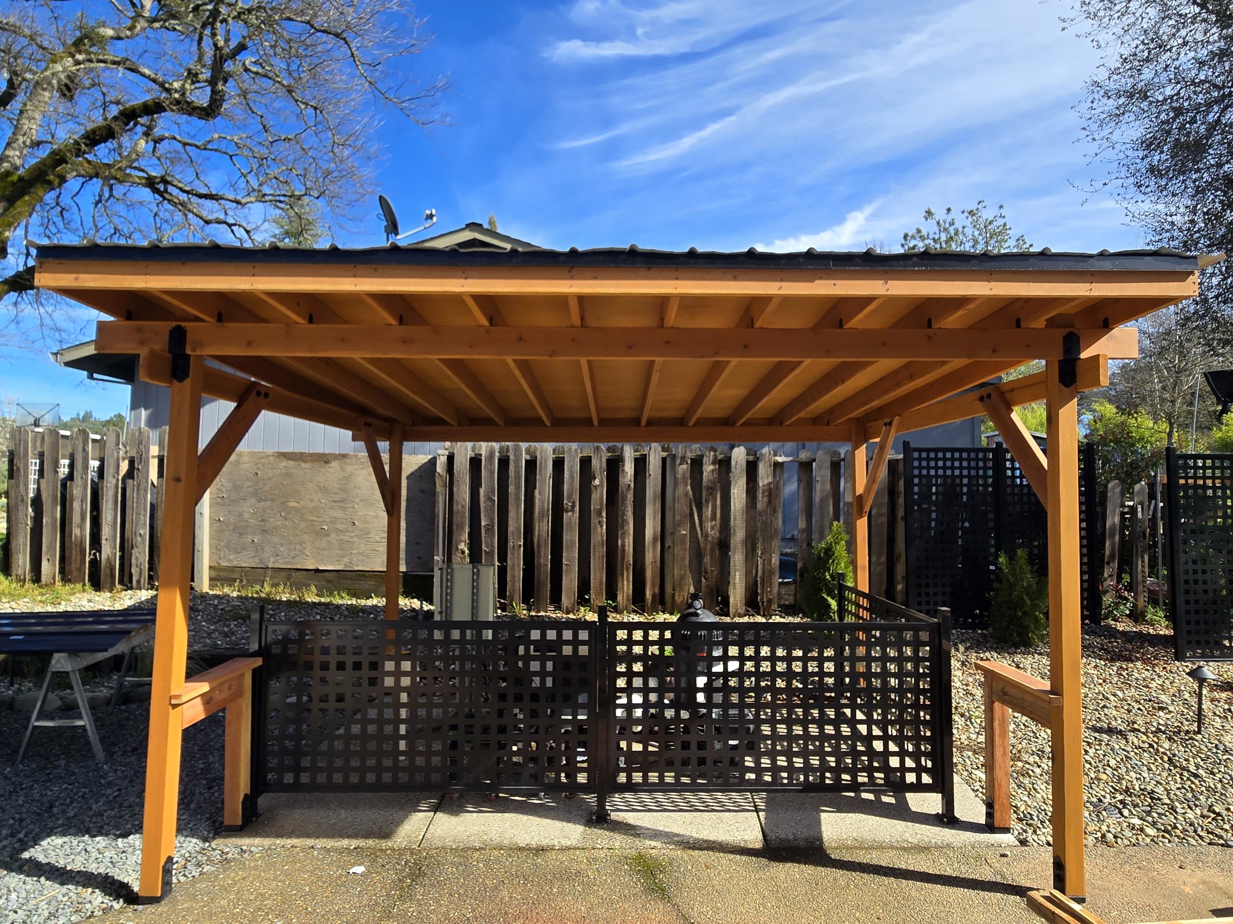 Open-sided wood shade structure with a metal roof and black lattice screening, blue sky behind